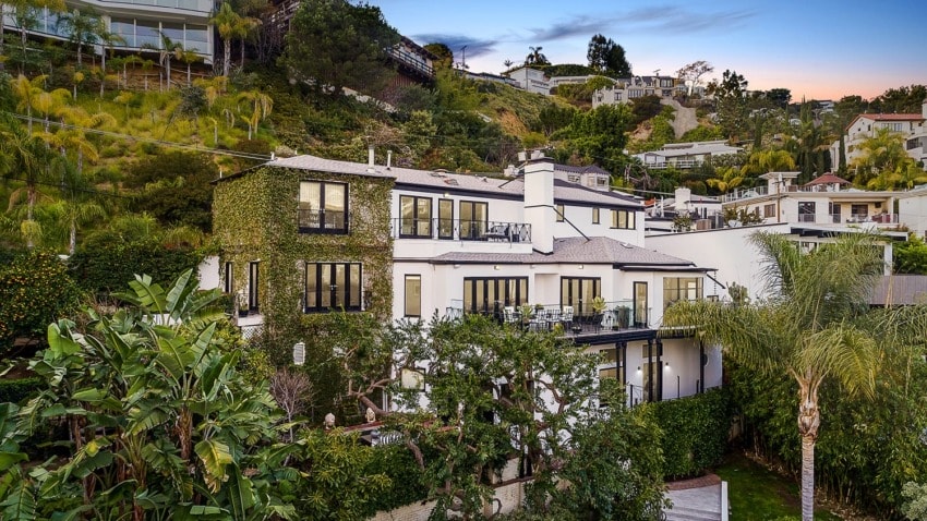 Luxury hillside home covered in ivy with large windows and spacious balconies surrounded by lush greenery at sunset.