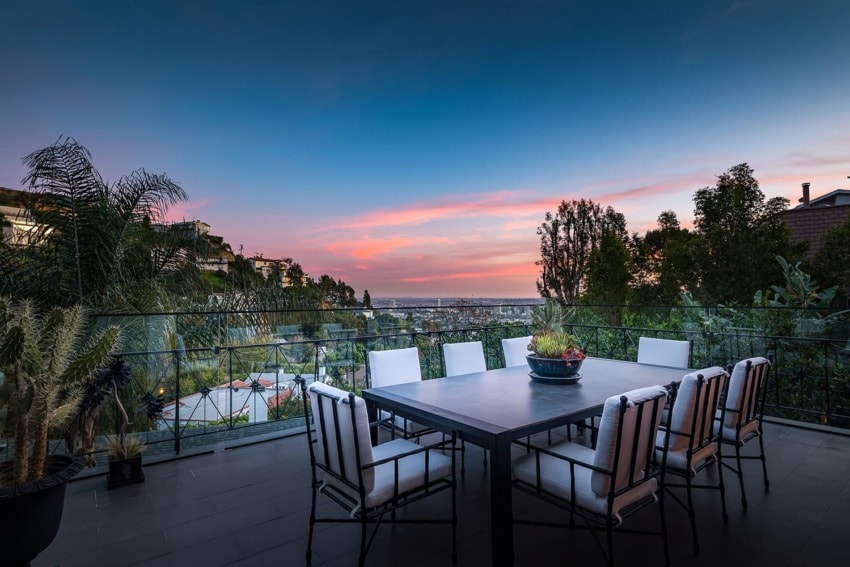 Outdoor patio dining area with white chairs, a large table, and a view of the city skyline at sunset.