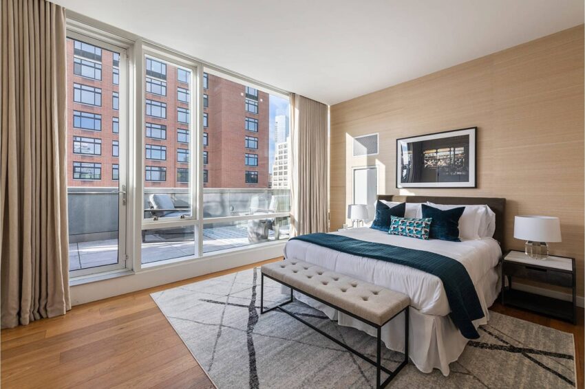 Modern bedroom with large windows, a cozy bed, beige walls, abstract art, and city view in the background.