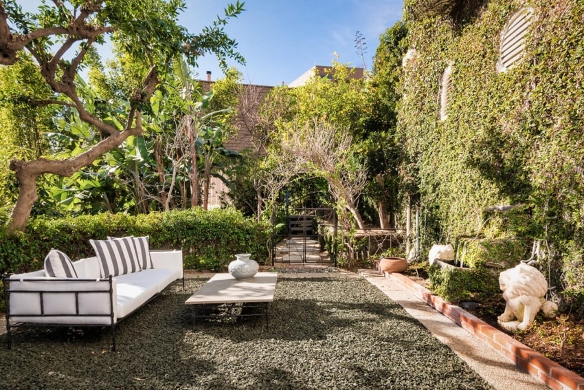 Outdoor garden with cushioned seating, lush green vines, trees, and decorative statues creating a serene and inviting atmosphere