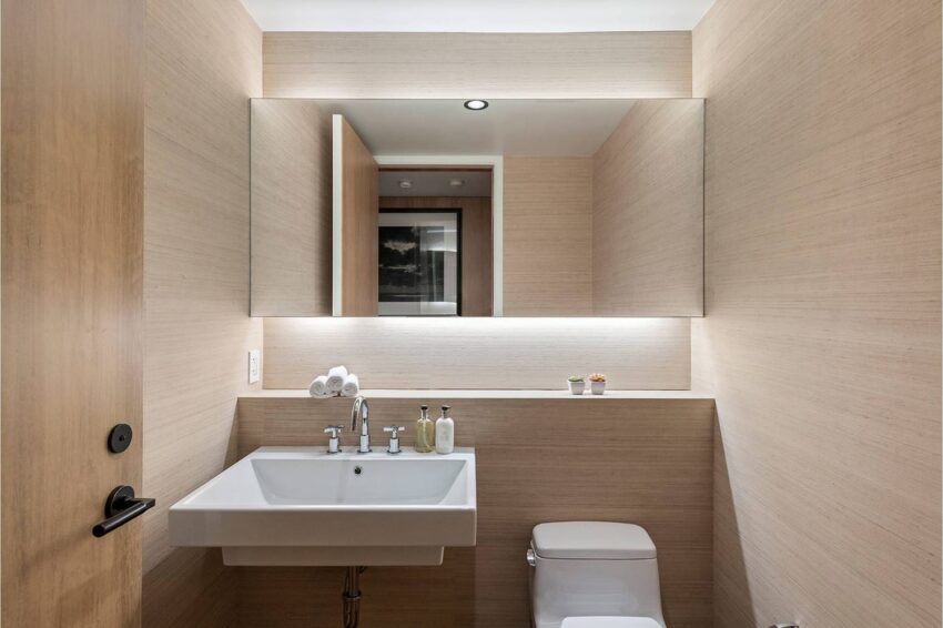 Modern bathroom with beige walls, a white sink, and a toilet. Large mirror reflecting lights. Plants on sink ledge.