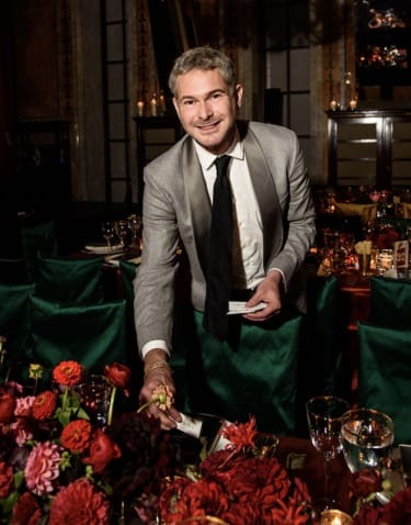 Person in a suit placing objects on a table with flowers and glassware in a dimly lit room.
