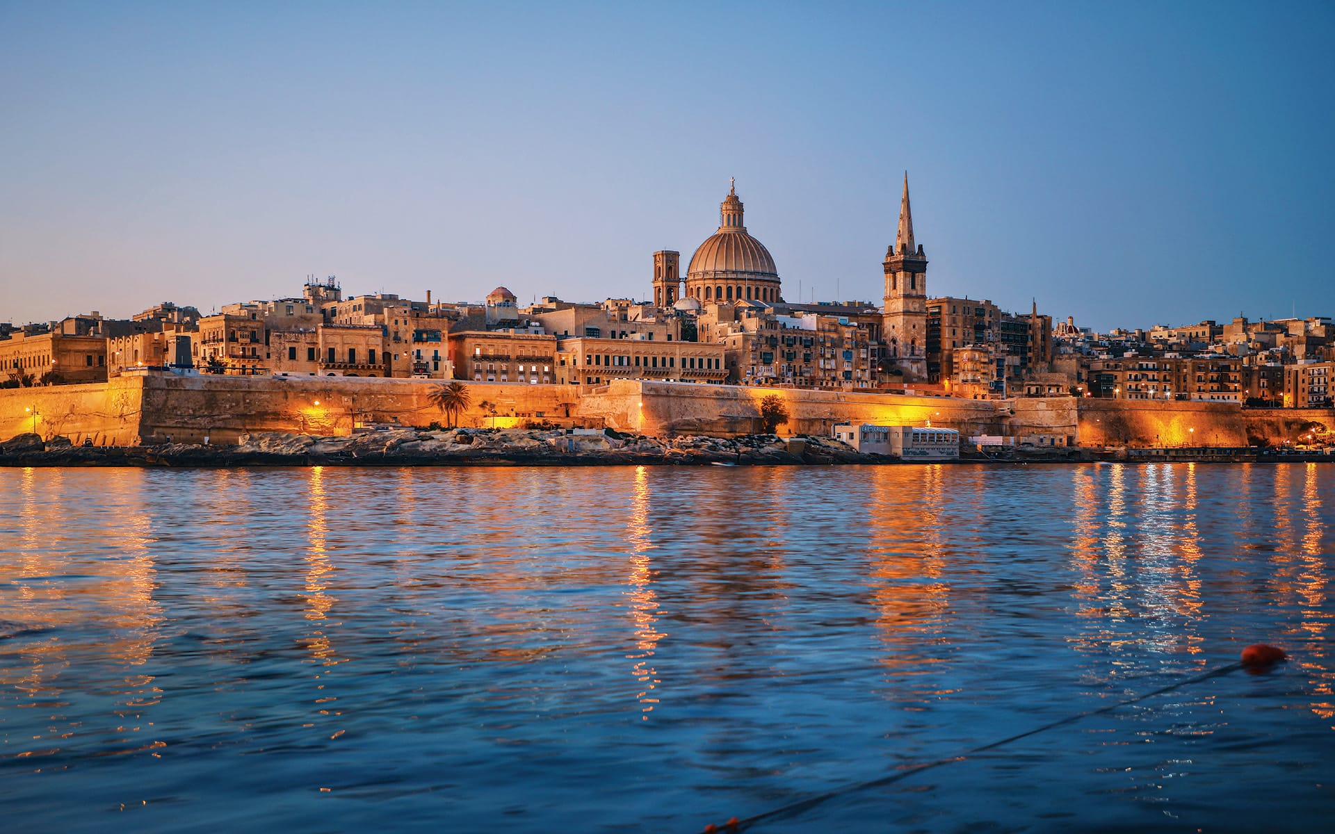 Skyline of Valletta, Malta at sunset with illuminated historic buildings reflecting on calm water.