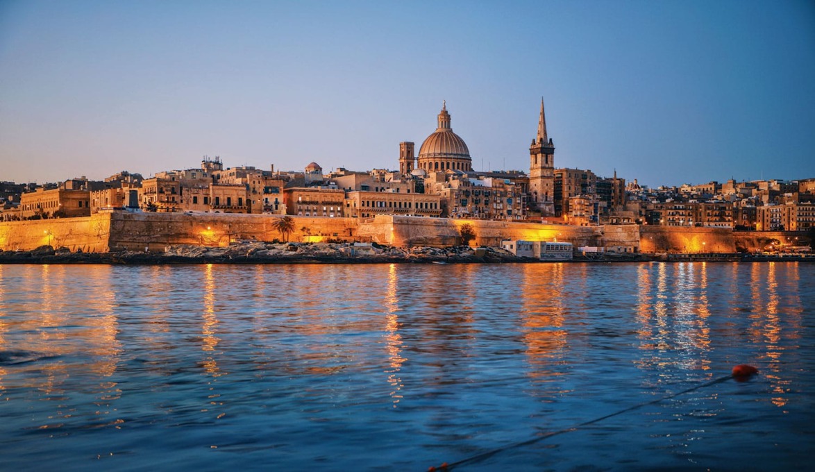Cityscape of Valletta, Malta at dusk with illuminated historic buildings and serene waterfront reflections.