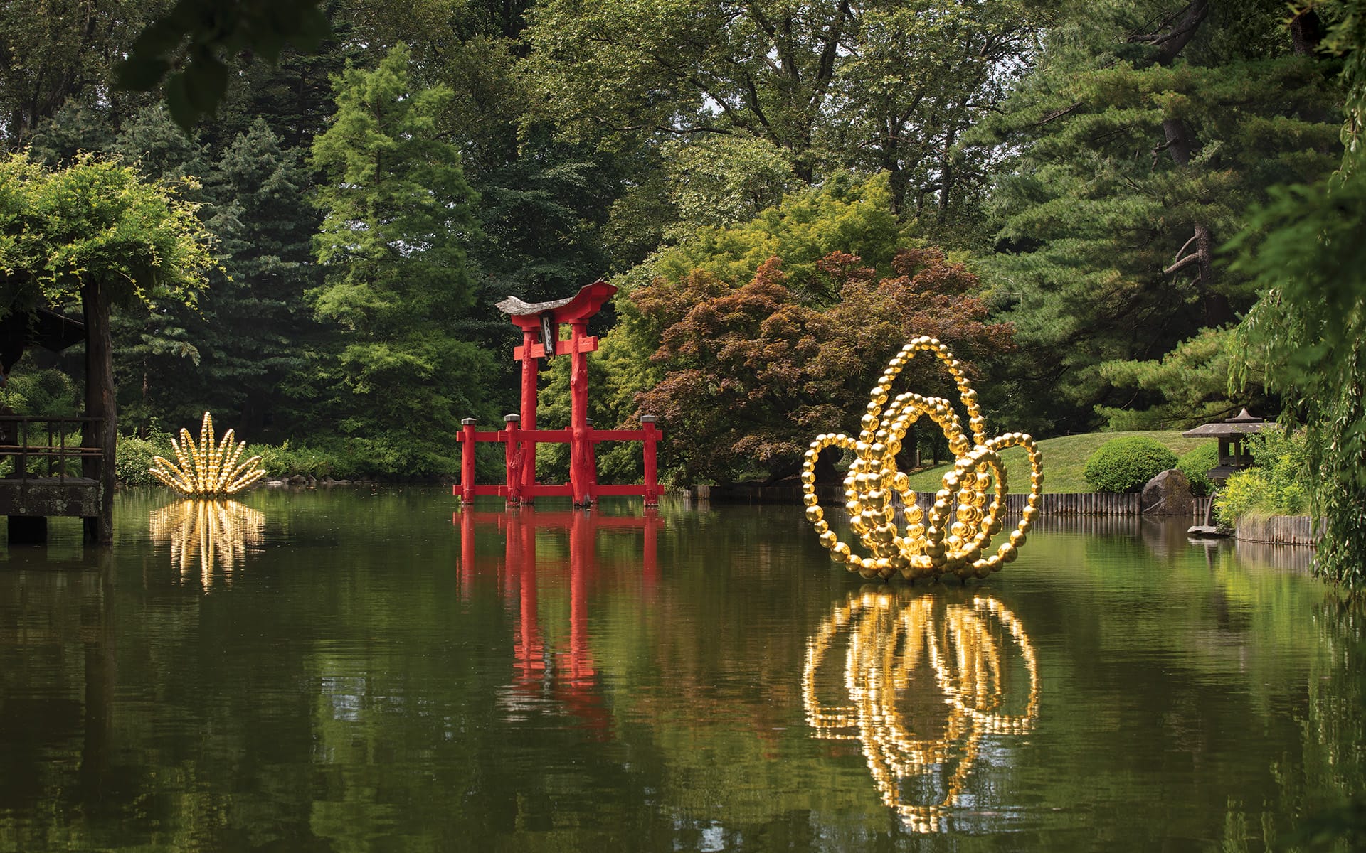 A serene pond with a red torii gate and illuminated lotus-shaped sculptures surrounded by lush greenery.