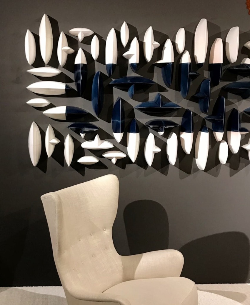 Modern art installation with abstract shapes on a dark wall behind a cream-colored upholstered chair.