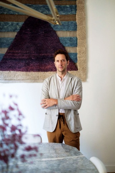 Person standing in front of a geometric tapestry, arms crossed, wearing a light gray blazer and brown pants.