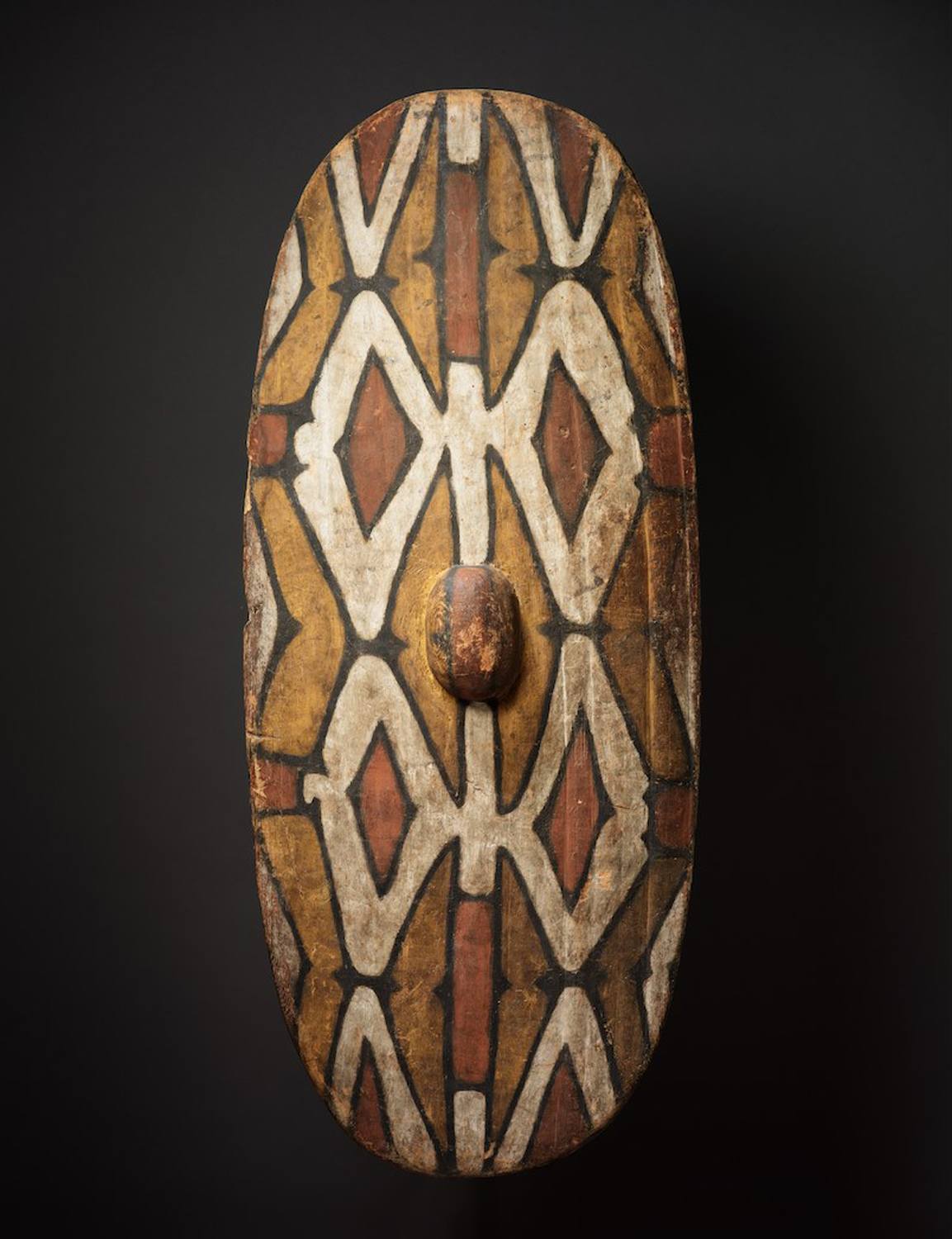 Wooden shield with geometric patterns in shades of brown, beige, and red, featuring an oval central boss.