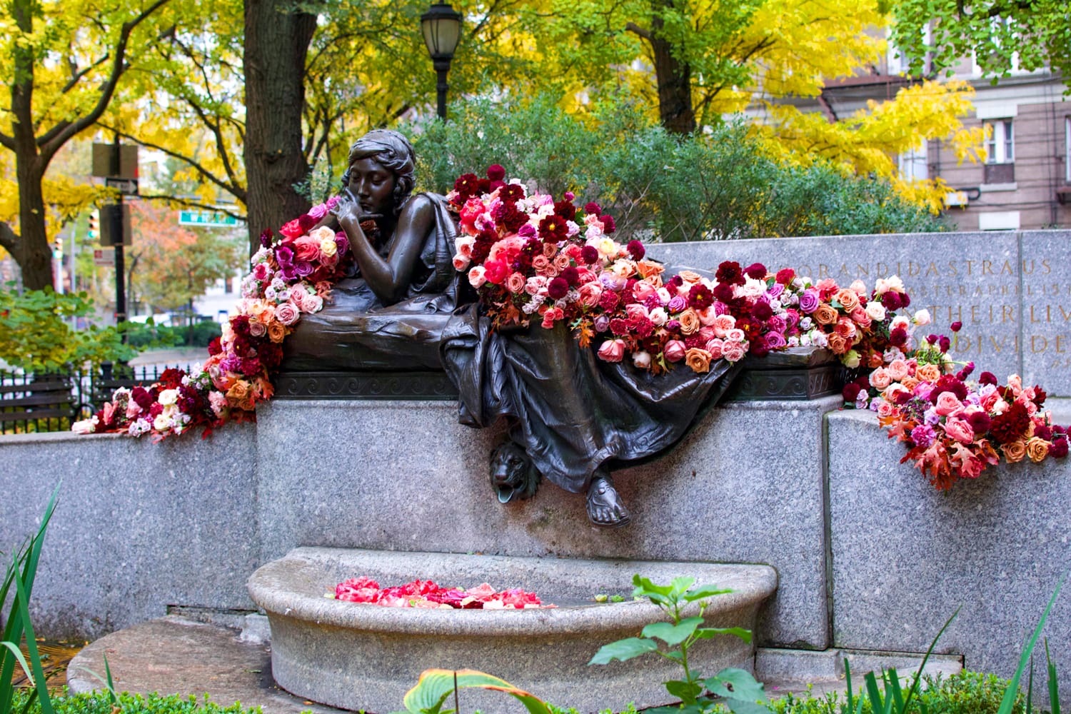 Statue of a seated figure adorned with red and pink flowers, surrounded by lush trees and vibrant autumn foliage.