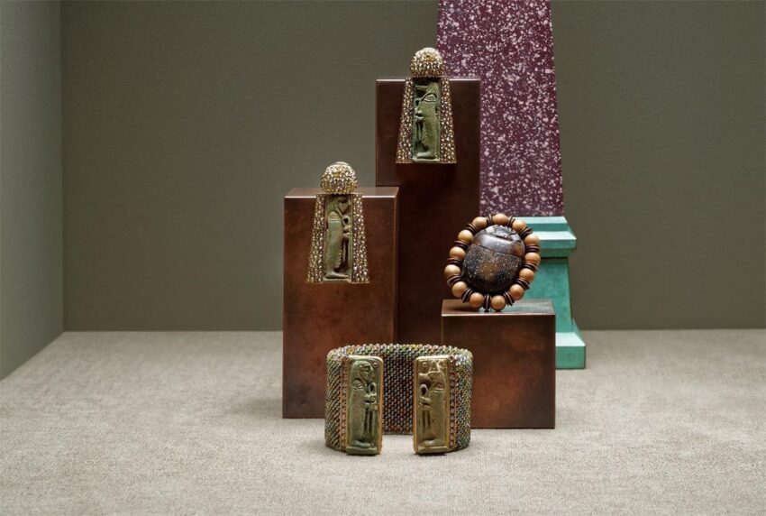 Display of ornate bronze bracelets and a wooden bead bracelet on geometric stands against a textured backdrop.