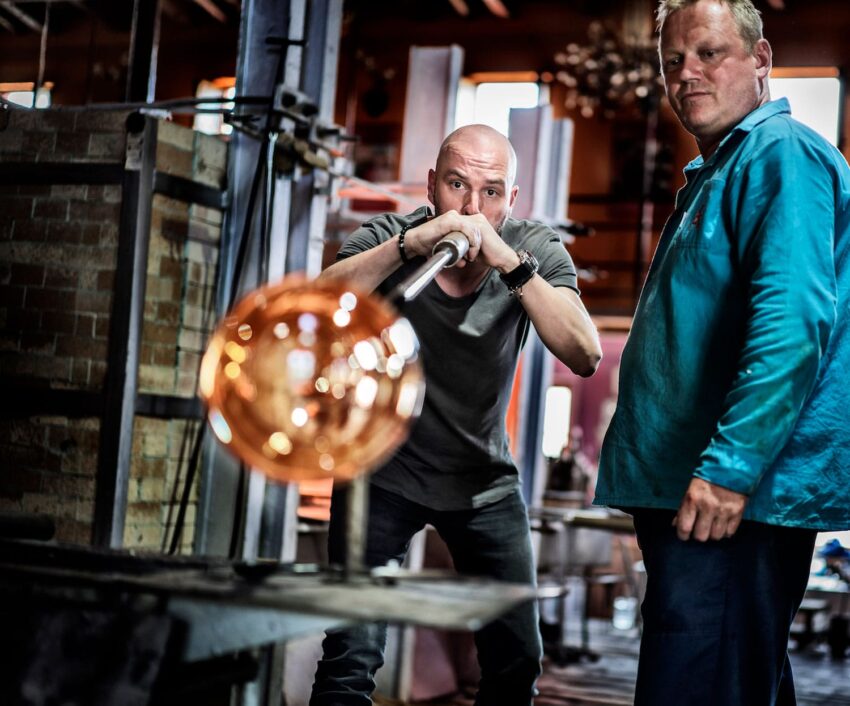 Glassblower shaping molten glass while a man observes in a workshop, showcasing traditional glassblowing techniques.