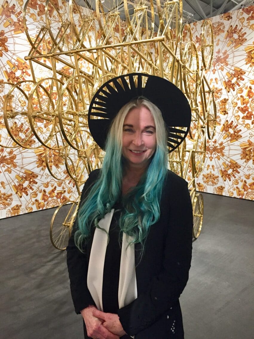 Person with long turquoise hair and a black hat in front of intricate golden art installation with wheels and floral patterns.