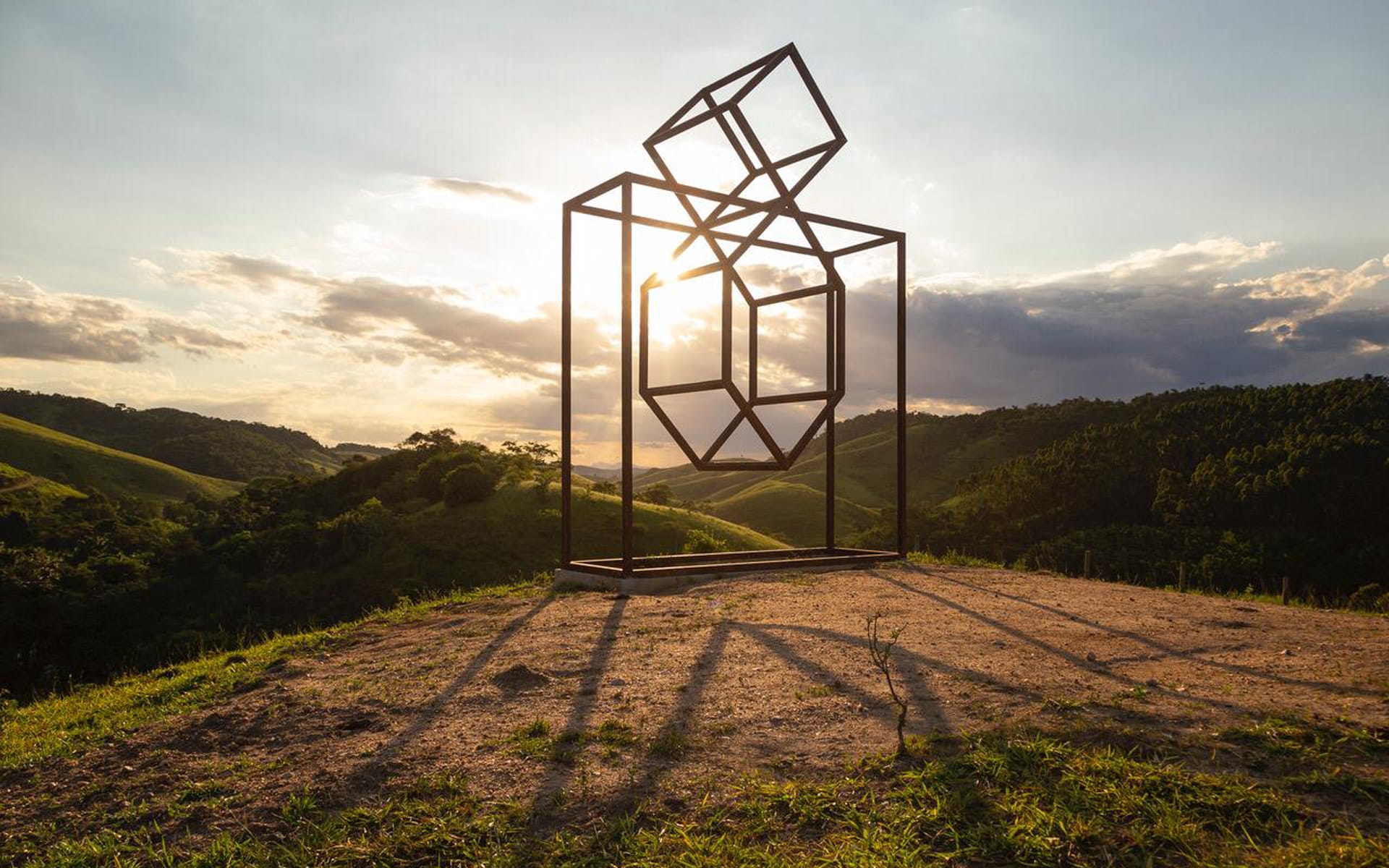 Geometric metal sculpture on a hilltop at sunset, casting long shadows over the grass with a scenic valley in the background.