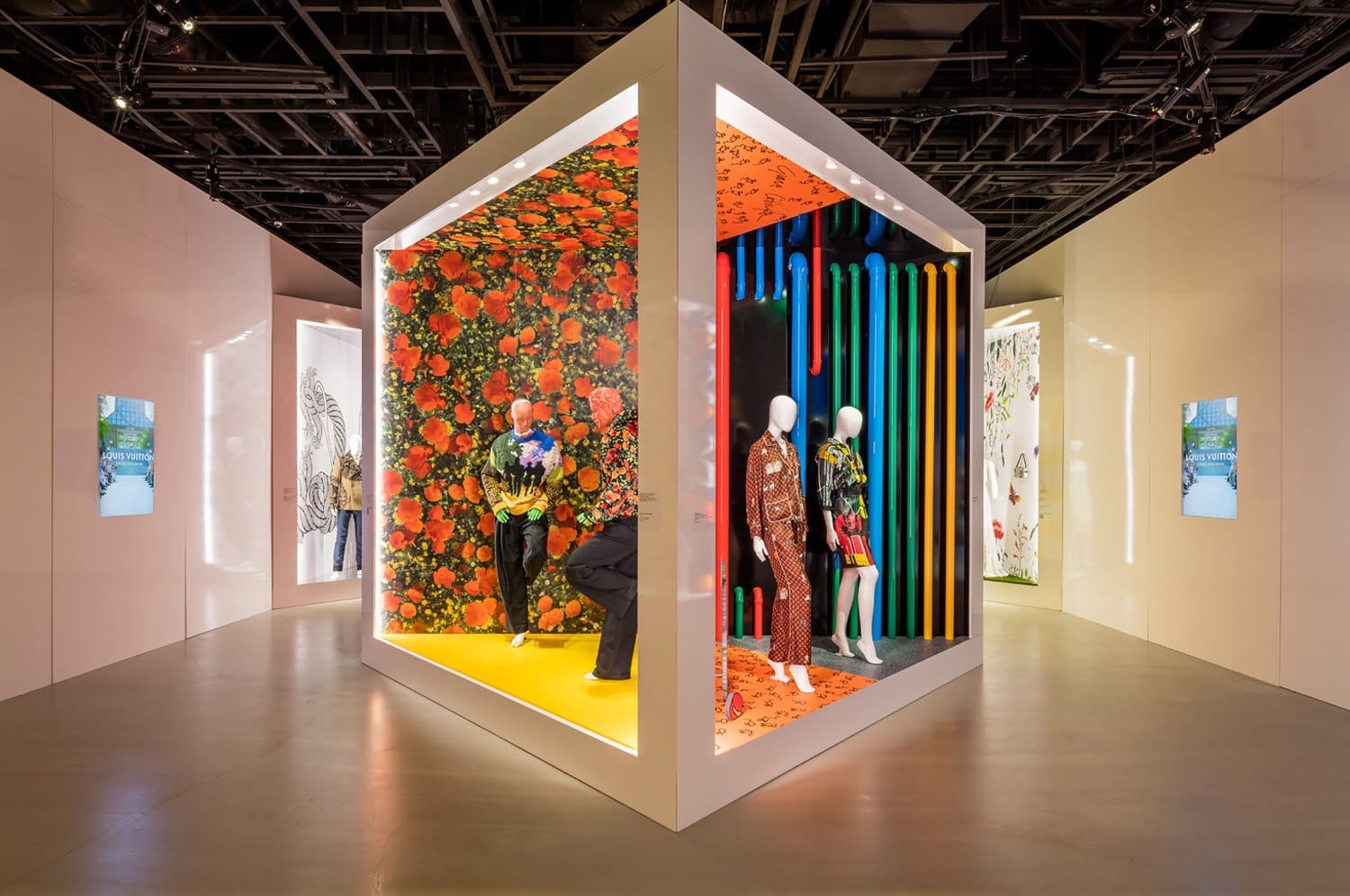 Colorful art installation with mannequin displays inside a cube, featuring vibrant floral and striped patterns on walls.