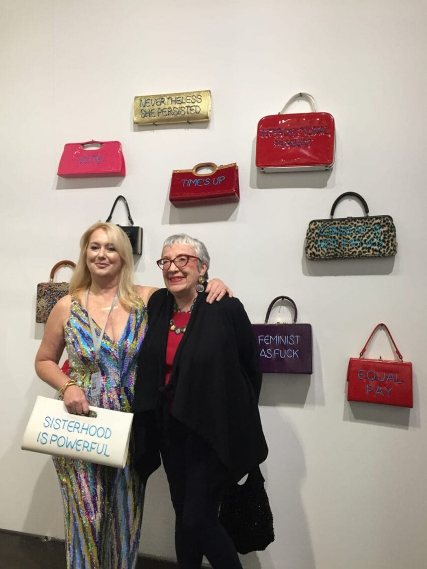 Two women standing in front of a wall displaying colorful purses with empowering slogans.