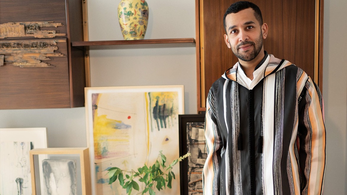 Man in a striped robe stands in a room with abstract art and a decorative vase on shelves.