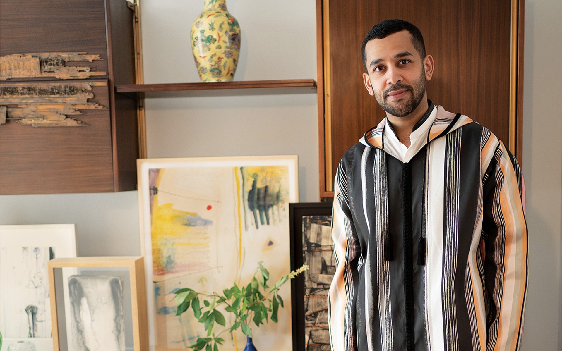 Man in a striped robe stands in a room with abstract art and a decorative vase on shelves.