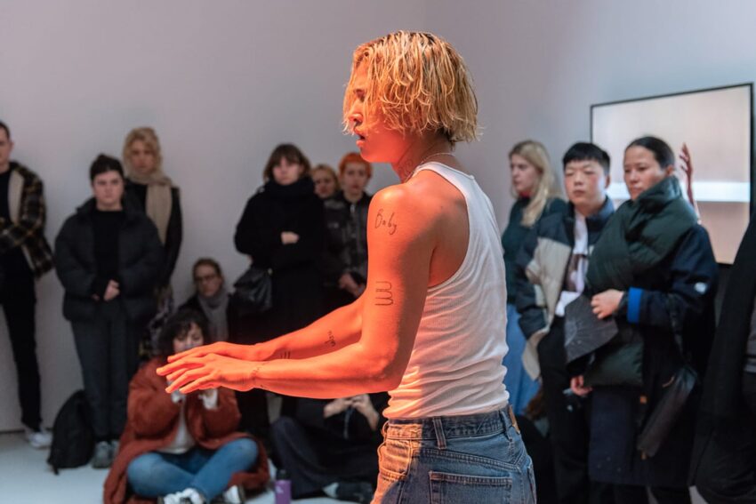 Person with blond hair performing in front of seated and standing audience in a casual indoor setting.