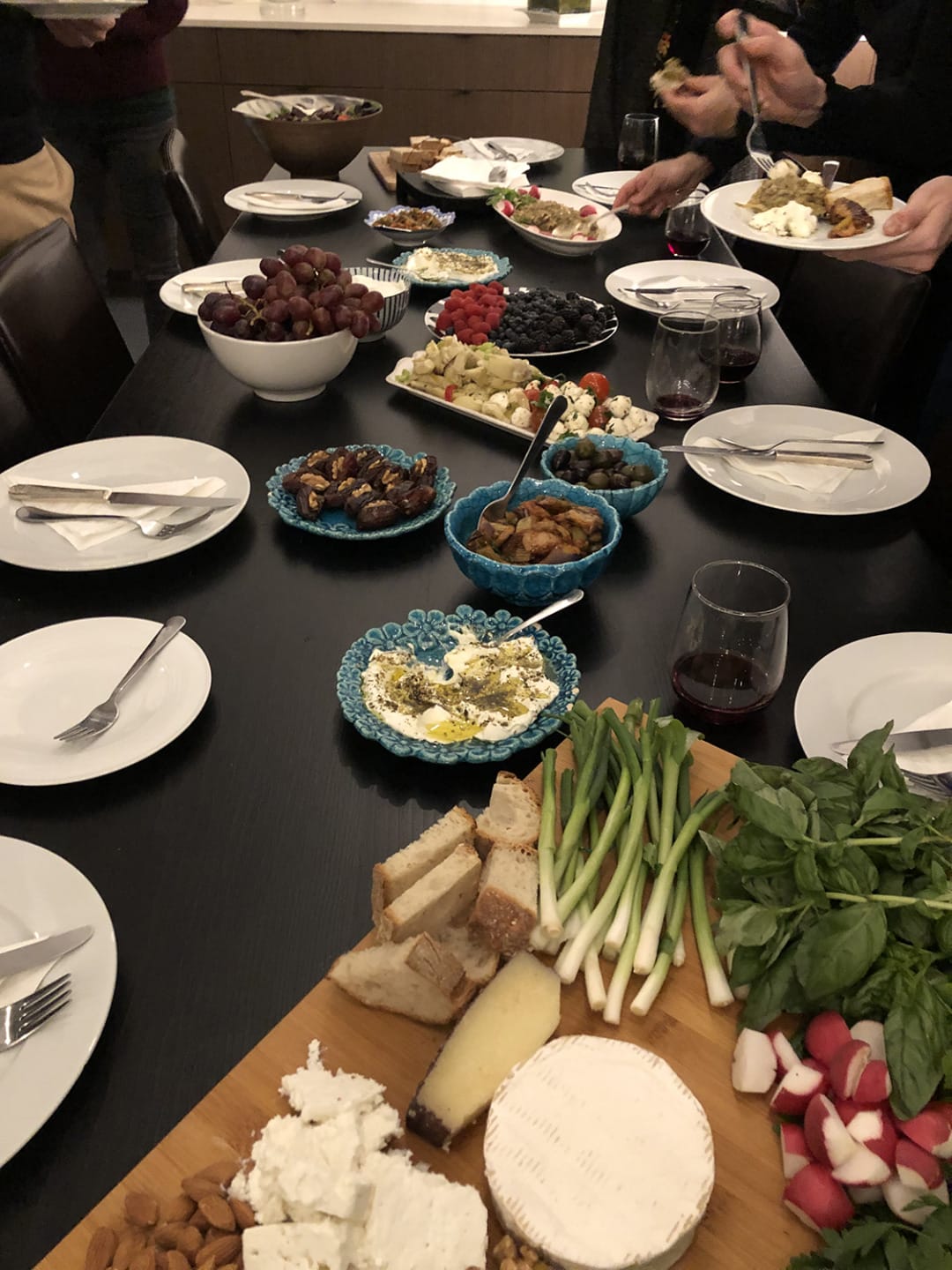 Assorted appetizers on a dining table including cheese, bread, vegetables, and people serving themselves.