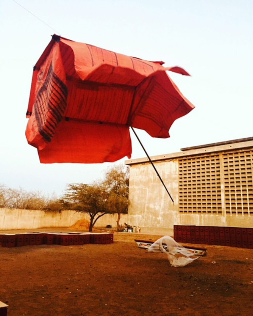 Red fabric kite suspended in the air against a clear sky, with a building and tree in the background.