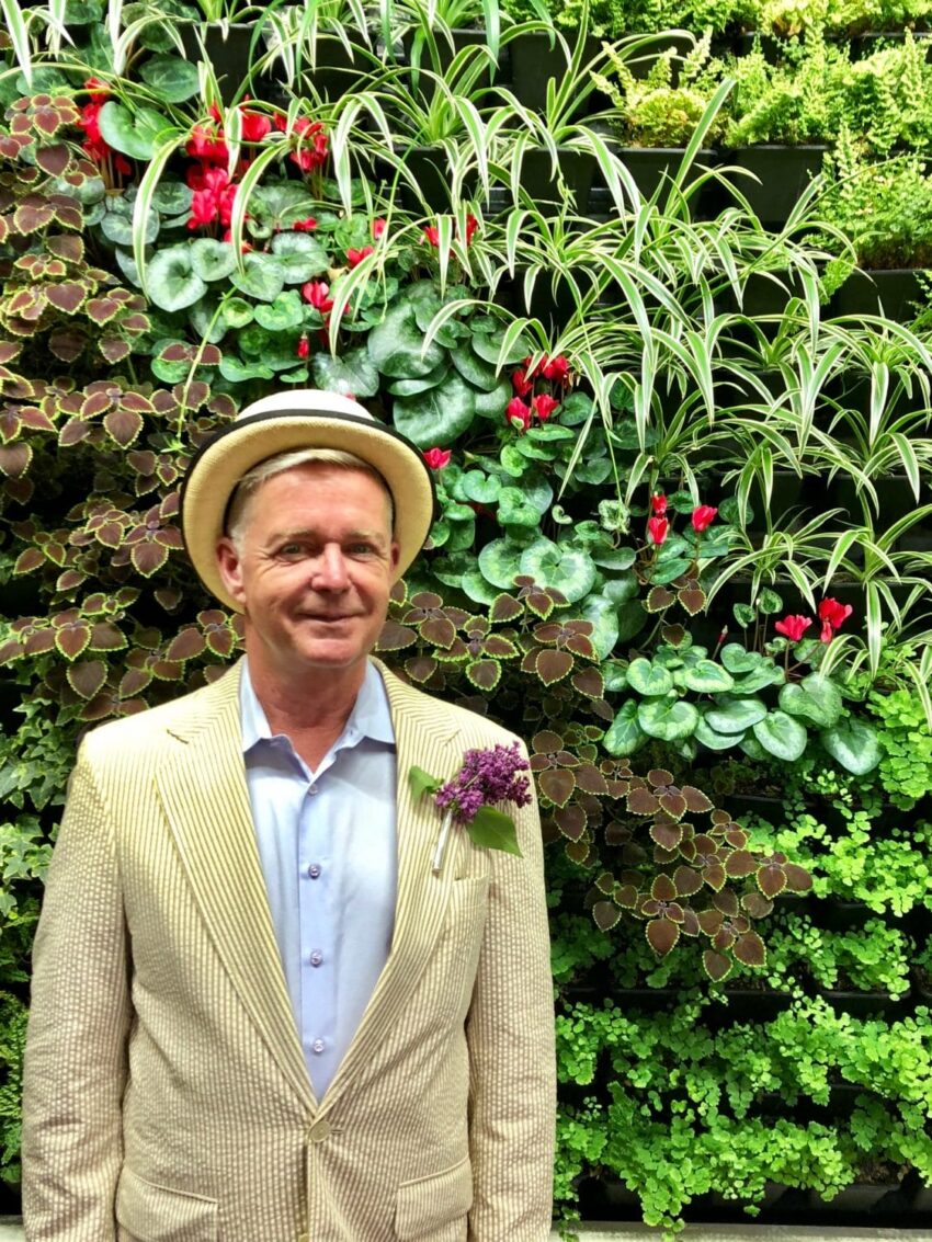 Man in a hat and suit stands in front of a lush vertical garden with diverse green and red plants.