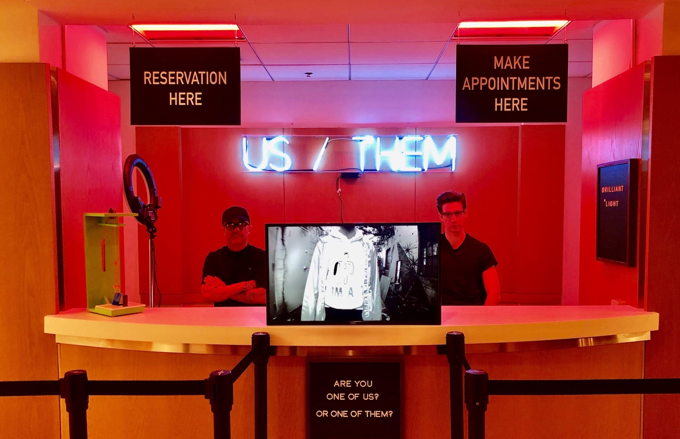 Art installation featuring a neon sign reading "US / THEM" with two people standing behind a counter in red lighting.