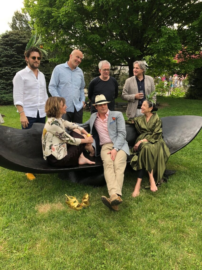 Group of people outdoors sitting and standing around a black sculpture, with trees and grass in the background.