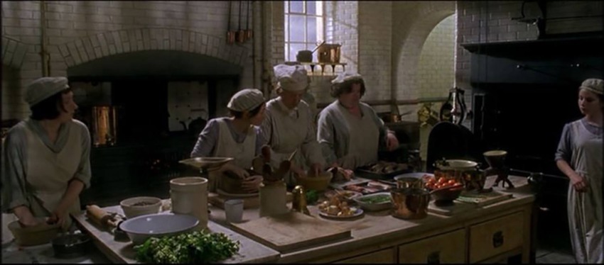 Several people in period attire working in a vintage kitchen with various cookware and ingredients on a wooden table.