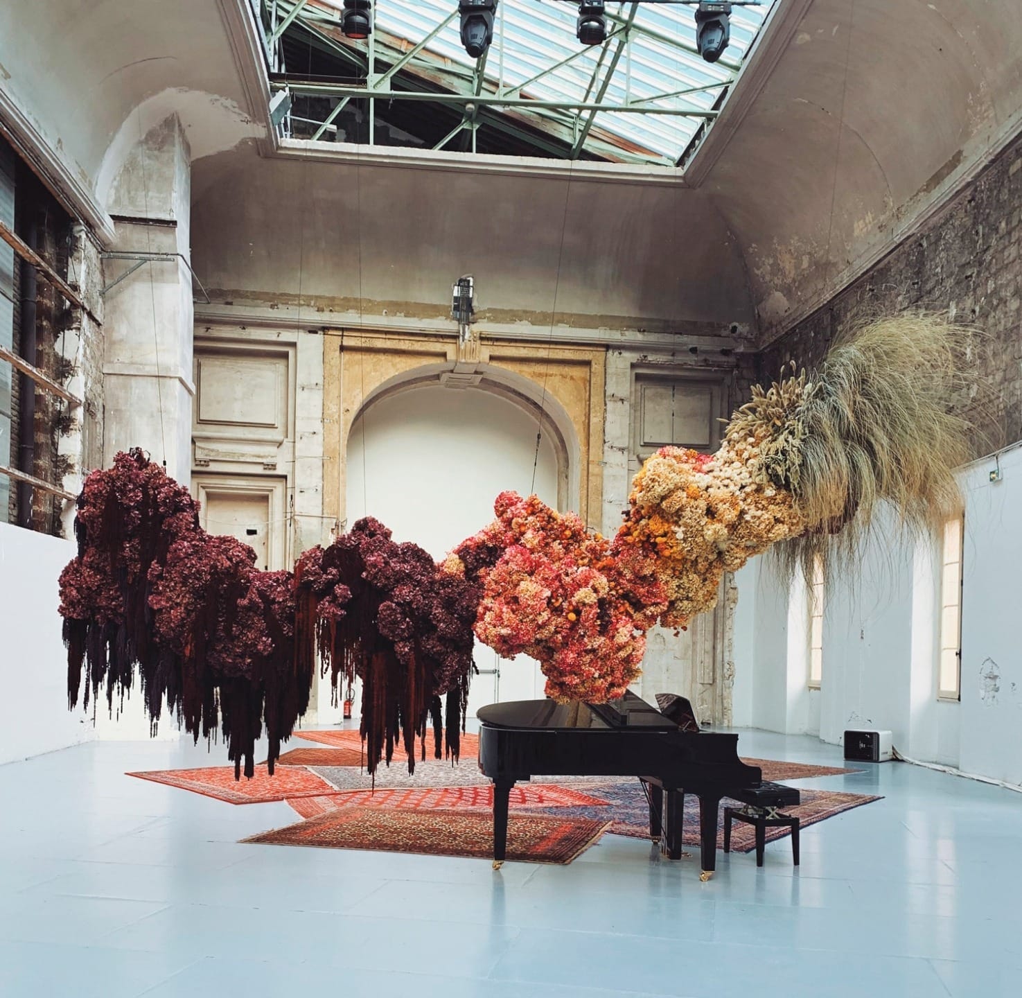 Art installation with vibrant floral sculptures suspended in a spacious gallery, featuring a grand piano on the floor.