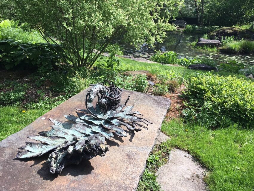 Bronze leaf sculpture on a stone slab in a lush garden with a pond in the background.
