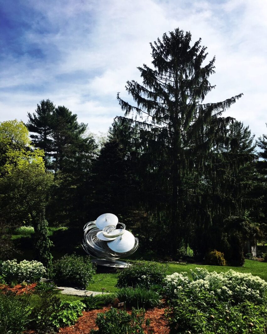 Sculptural art piece in a garden with tall trees and a clear sky in the background.