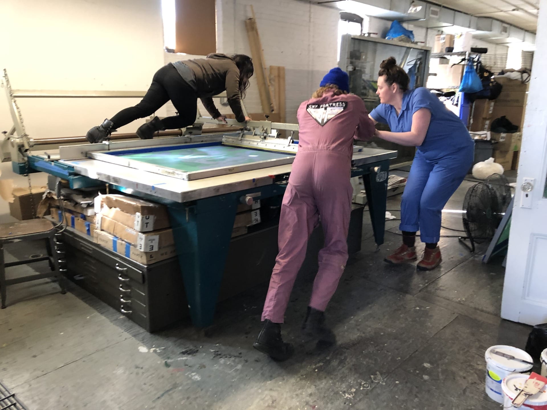 Three people in a workshop collaboratively working on a large screen printing table with supplies around them.