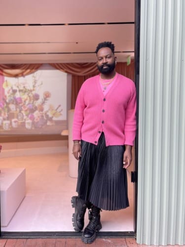 Person in a pink cardigan and black pleated skirt standing by an open door with a floral arrangement in the background.