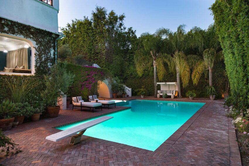 Luxurious backyard with illuminated swimming pool, surrounded by lush greenery and comfortable lounge chairs on a brick patio.