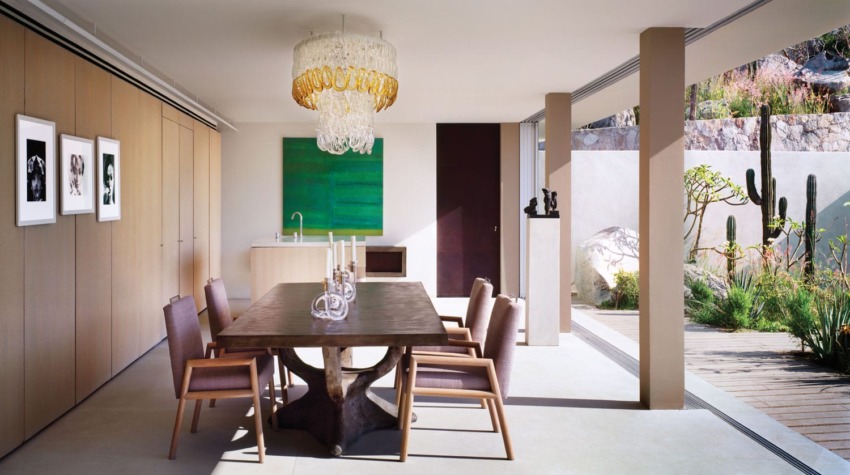 Modern dining room with wooden table, six chairs, chandelier, cacti, and large artwork on the wall in a bright space.