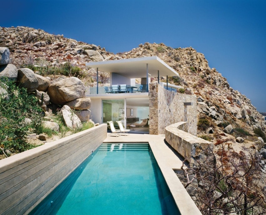 Modern cliffside house with a pool and outdoor lounge area, surrounded by rocky terrain under a clear blue sky.