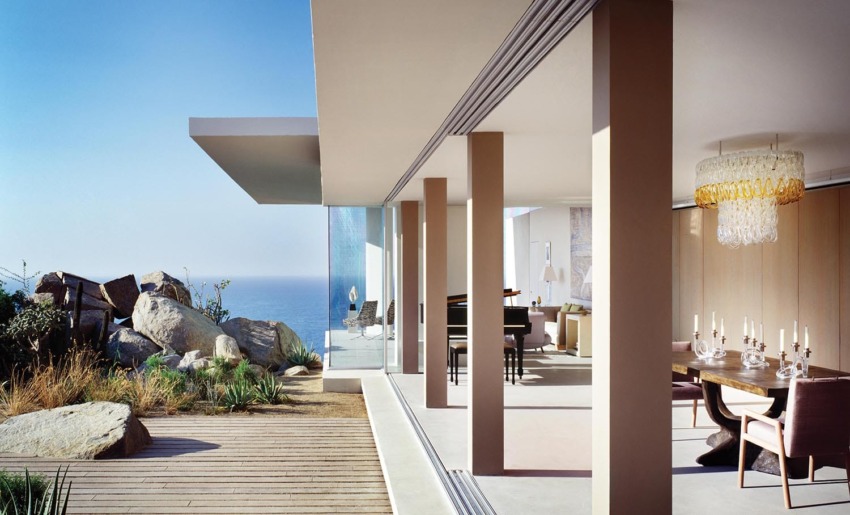 Modern coastal home with glass walls, ocean view, dining table, chandelier, and piano in an open living room.
