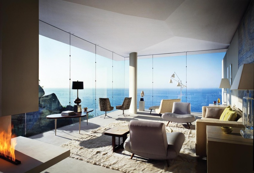 Modern living room with panoramic ocean views, featuring sleek furniture, large windows, and a cozy fireplace.
