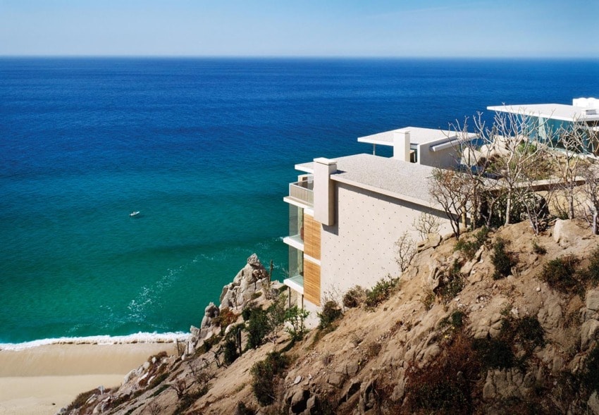 Modern cliffside house overlooking the ocean with clear blue water and sandy beach below.