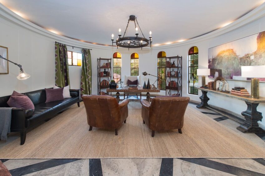 Spacious round living room with leather chairs, black sofa, chandelier, bookshelves, and large windows.