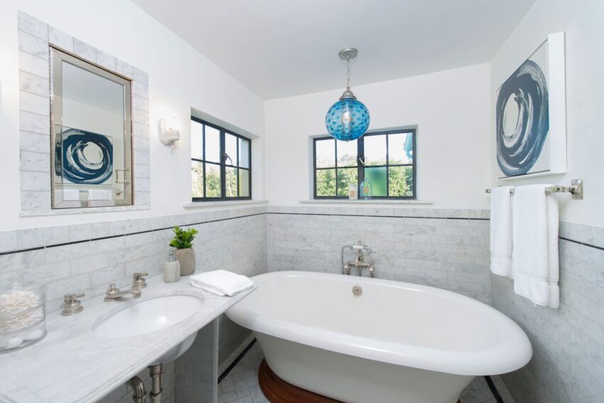 Modern bathroom with freestanding tub, marble countertop, blue accents, and natural light from large windows.