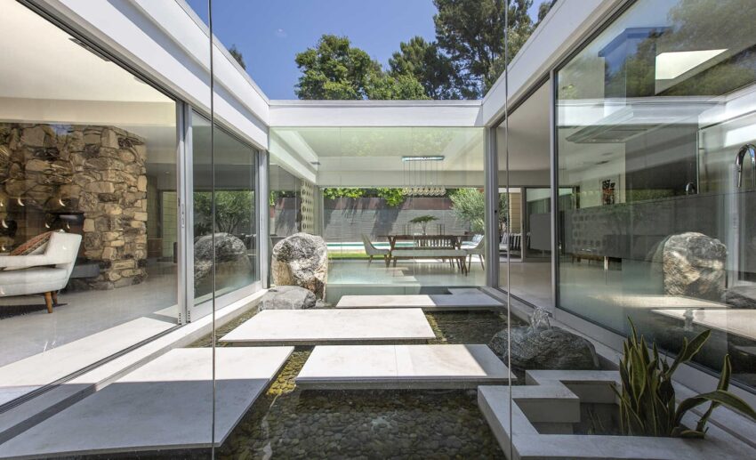 Modern glass-walled living area with indoor stone water feature and view of outdoor patio with greenery.