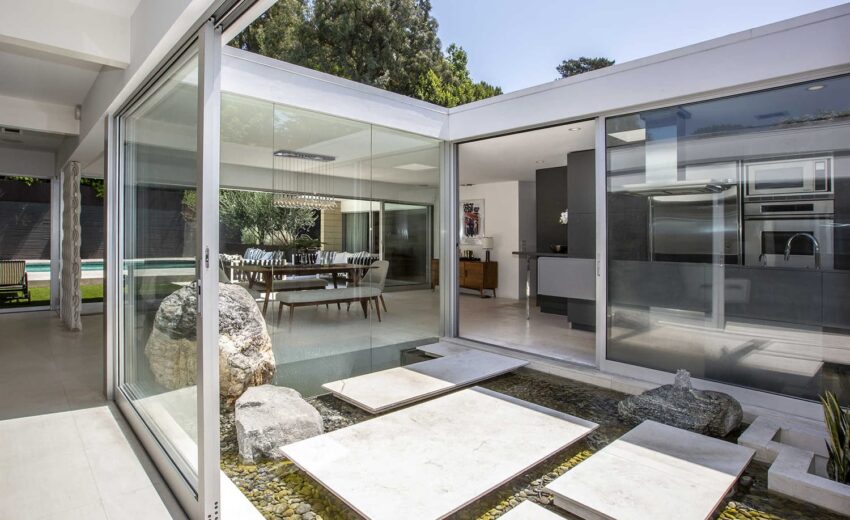 Modern open interior with glass walls, stone walkway over water feature, dining area, and kitchen in a minimalist home.