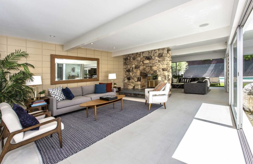 Spacious modern living room with large windows, gray sofa, wooden coffee table, and stone fireplace, opening to a patio.