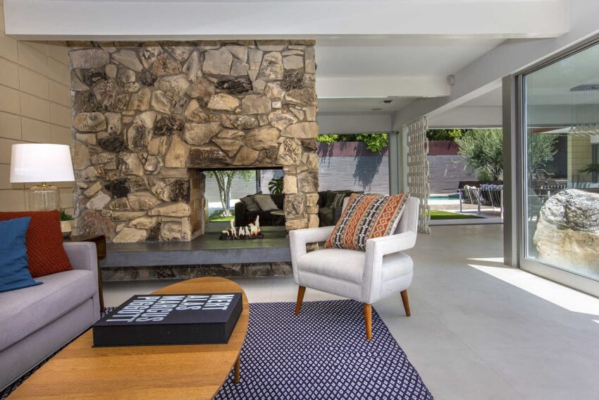 Modern living room with a stone fireplace, gray armchair, coffee table, and large windows overlooking a patio area.