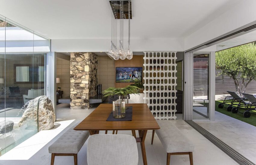 Modern dining area with a wooden table, bench seating, and a view of greenery and lounge chairs outside sliding glass doors.
