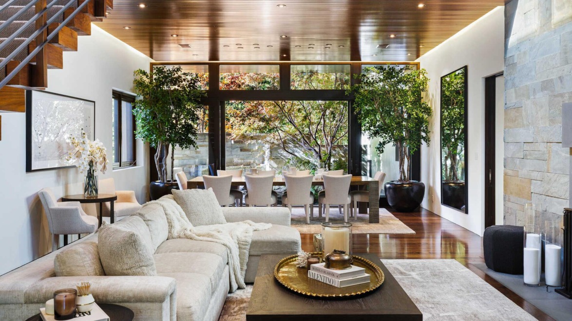 Modern living room with cozy sectional sofa, coffee table, and dining area in background, decorated with large indoor plants.