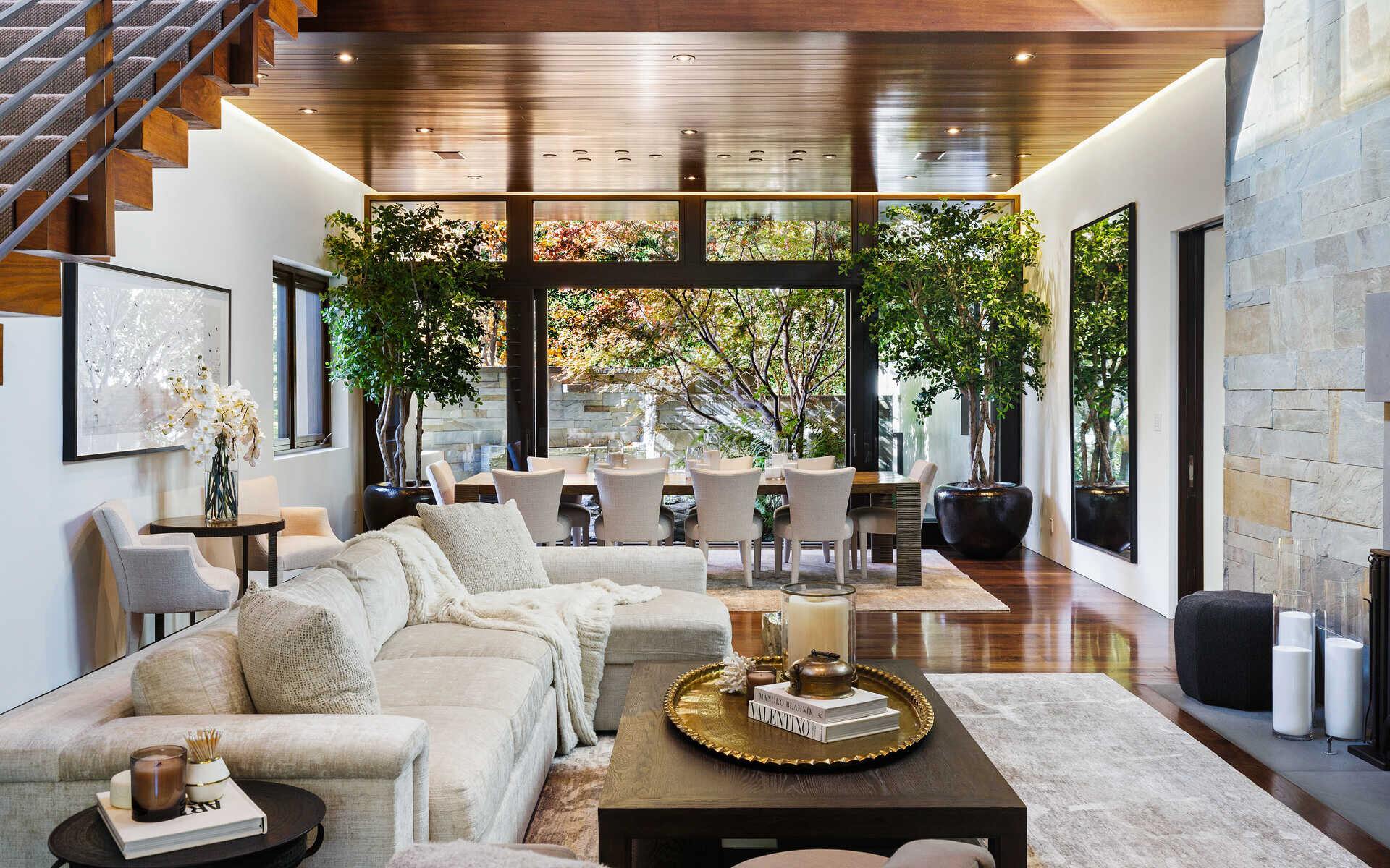 Modern living room with cozy sectional sofa, coffee table, and dining area in background, decorated with large indoor plants.