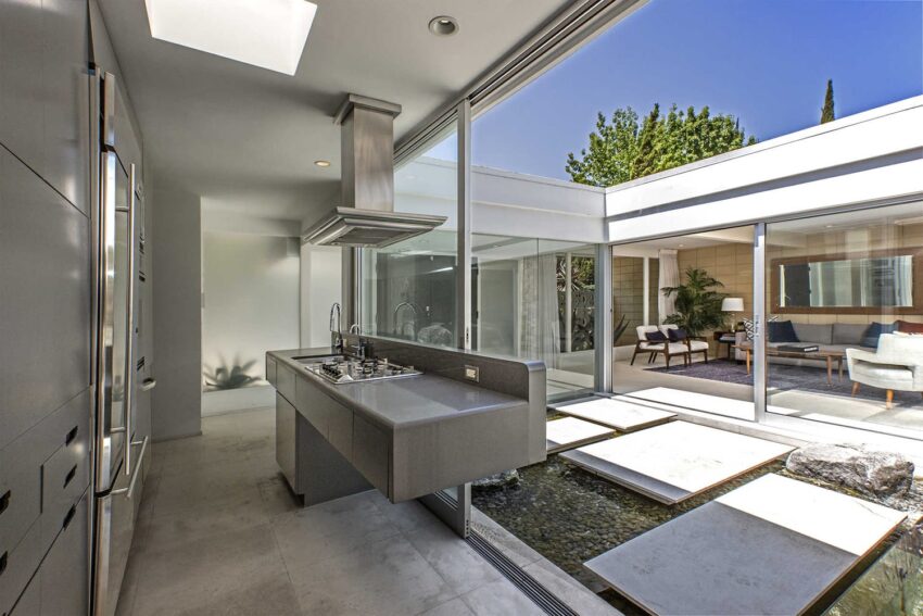 Modern kitchen with sleek gray island, large windows, and view of a courtyard with stepping stones and greenery.