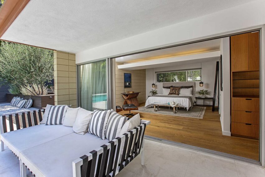 Modern bedroom opens onto patio with striped outdoor sofa, wood flooring, and natural decor elements.